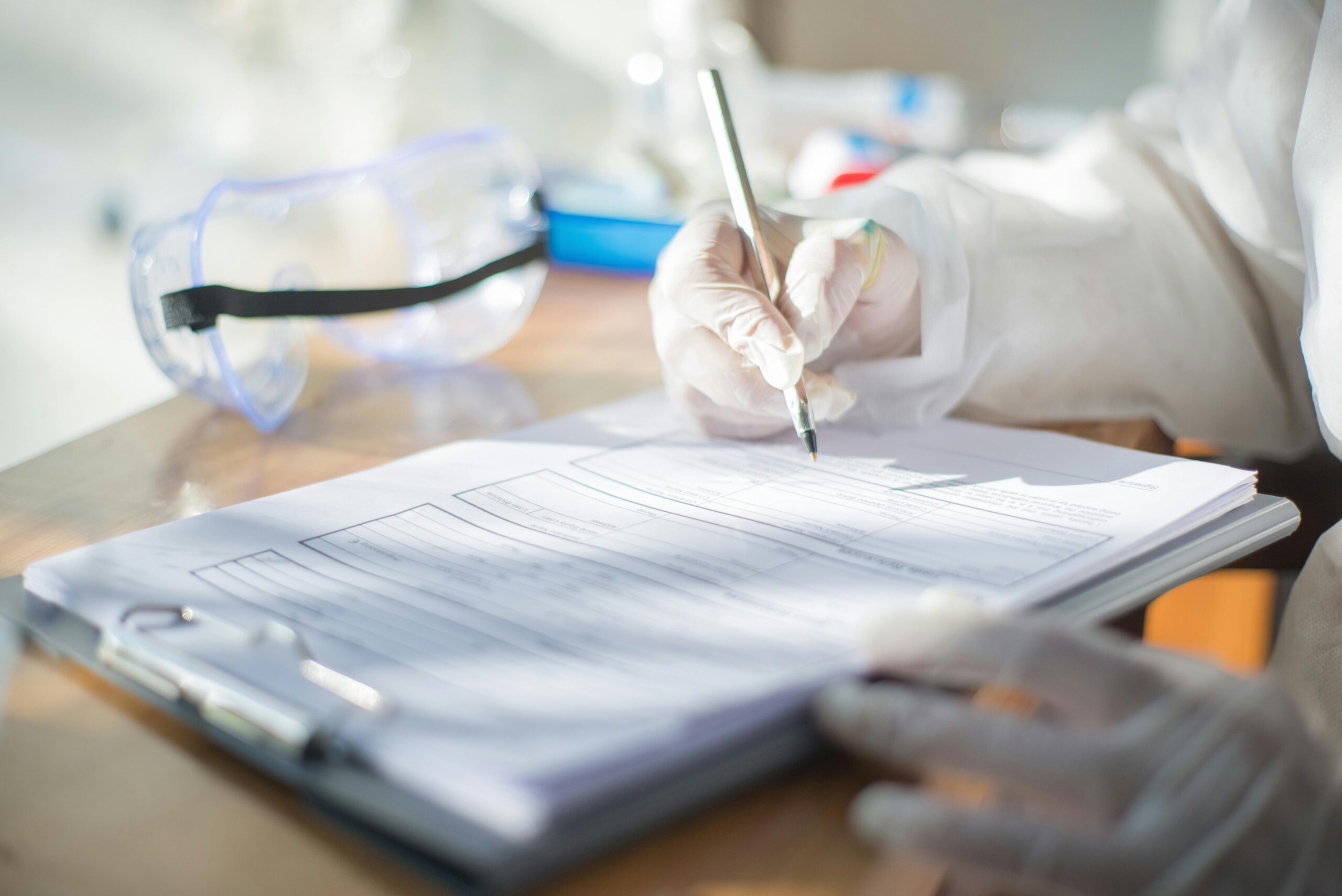 close up of a person in a lab coat and gloves filling out a checklist while sitting at a desk. safety goggles are also next to them.