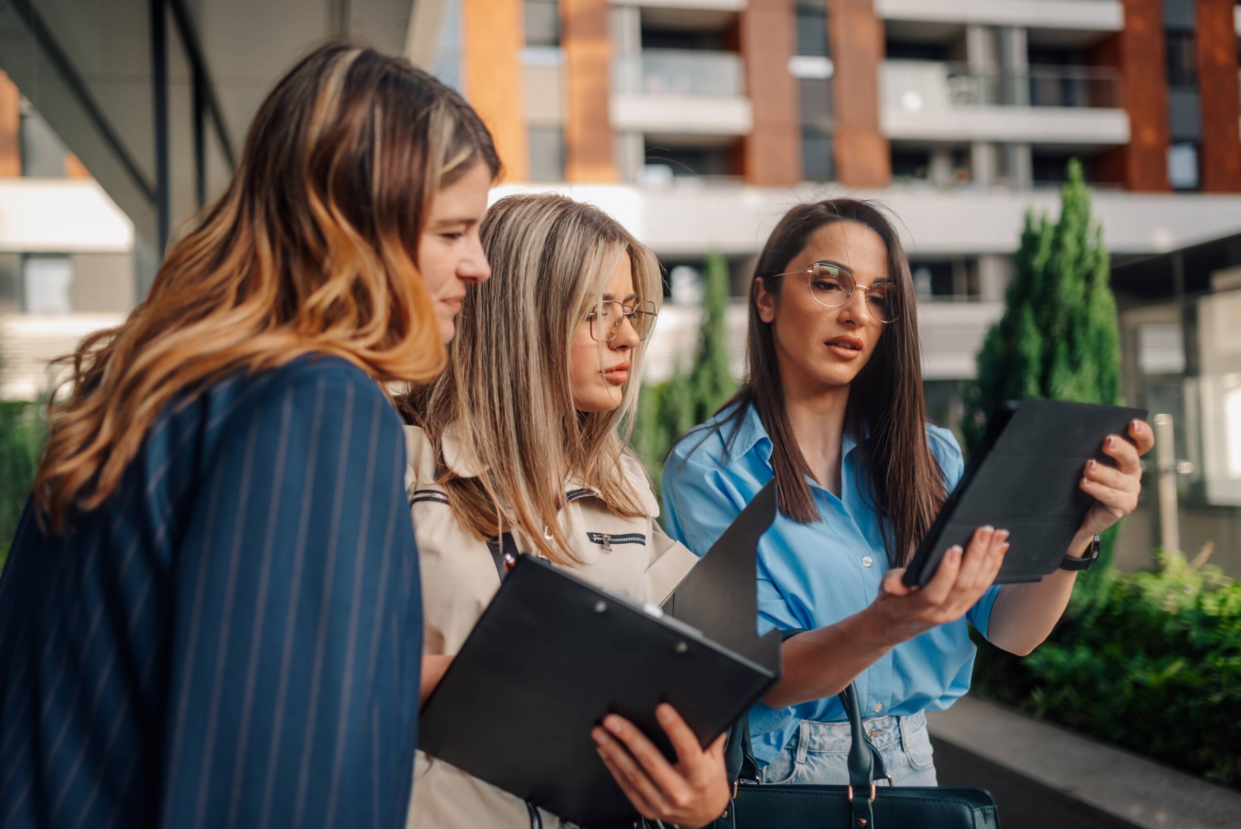 Three stylish young women stand outside an office building discussing business. Two of the women hold documents in their hands, and one woman holds a tablet. The women are dressed in fashionable clothing. The scene is outdoors, during daytime, under natural light.